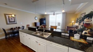A modern kitchen with granite countertops, a stainless steel sink, and white cabinetry. There is a dining area with a table and chairs, and a large window providing natural light.