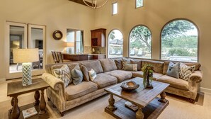 A spacious living room with a large sectional sofa, a wooden coffee table, and a chandelier. There are multiple windows providing natural light and views of the outdoors. - Cappella - Gorgeous 4 br Villa in PGA West with Pool/Spa + Golf Course Views #259222 (La Quinta)