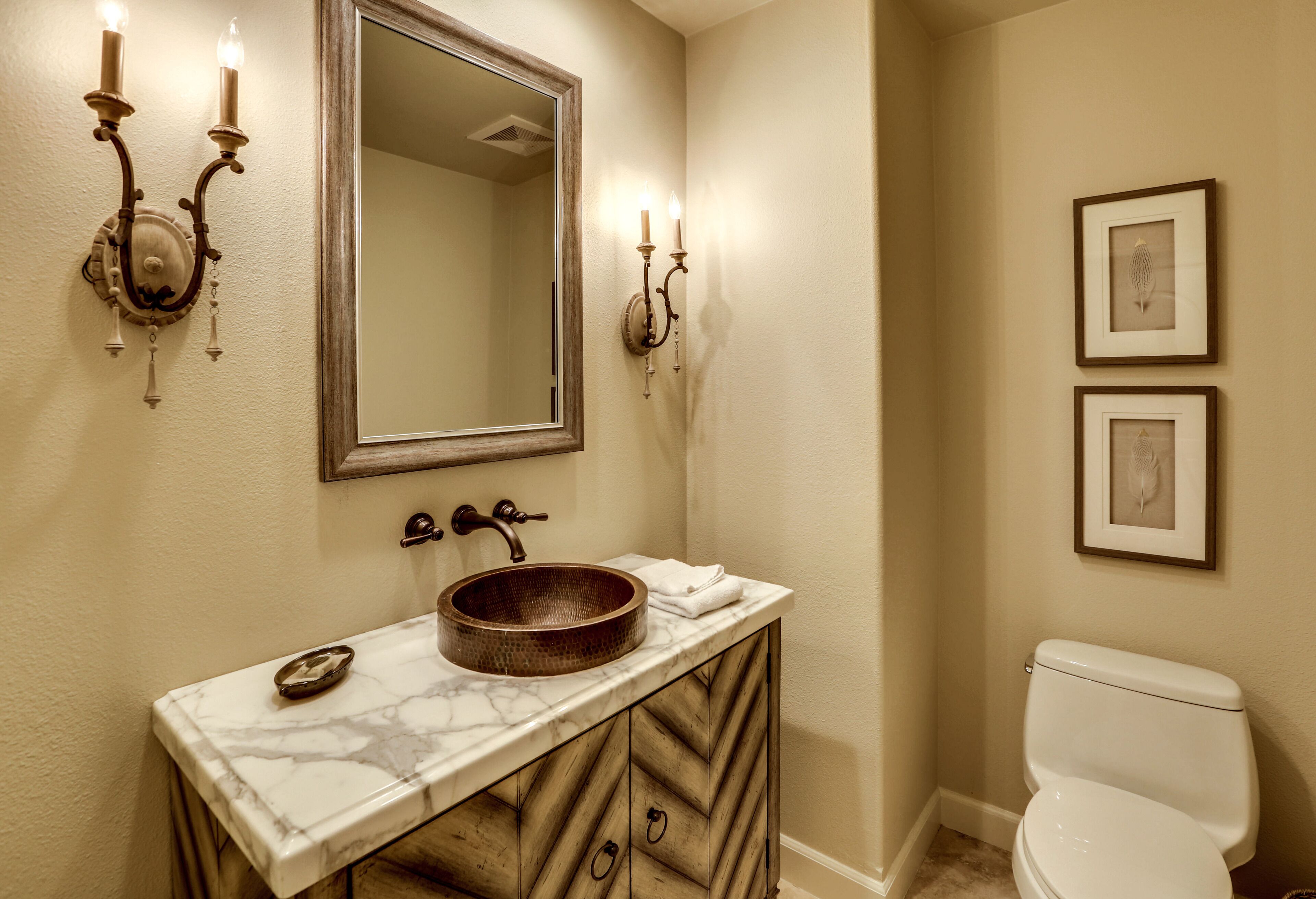 A bathroom with a marble sink, wooden vanity, a large mirror, wall sconces, and framed artwork.