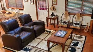A living room with a leather sofa, wooden chairs, a coffee table with a chessboard, a television, and a small table with a lamp and decorative items. - 3 Bedroom 2 bath Home, Easy Access, Covered Porch, Diligent Views & Internet (Maggie Valley)
