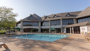 Un espace piscine avec des chaises longues et des parasols, un bâtiment doté de grandes fenêtres et de piliers en pierre, et un ciel dégagé.