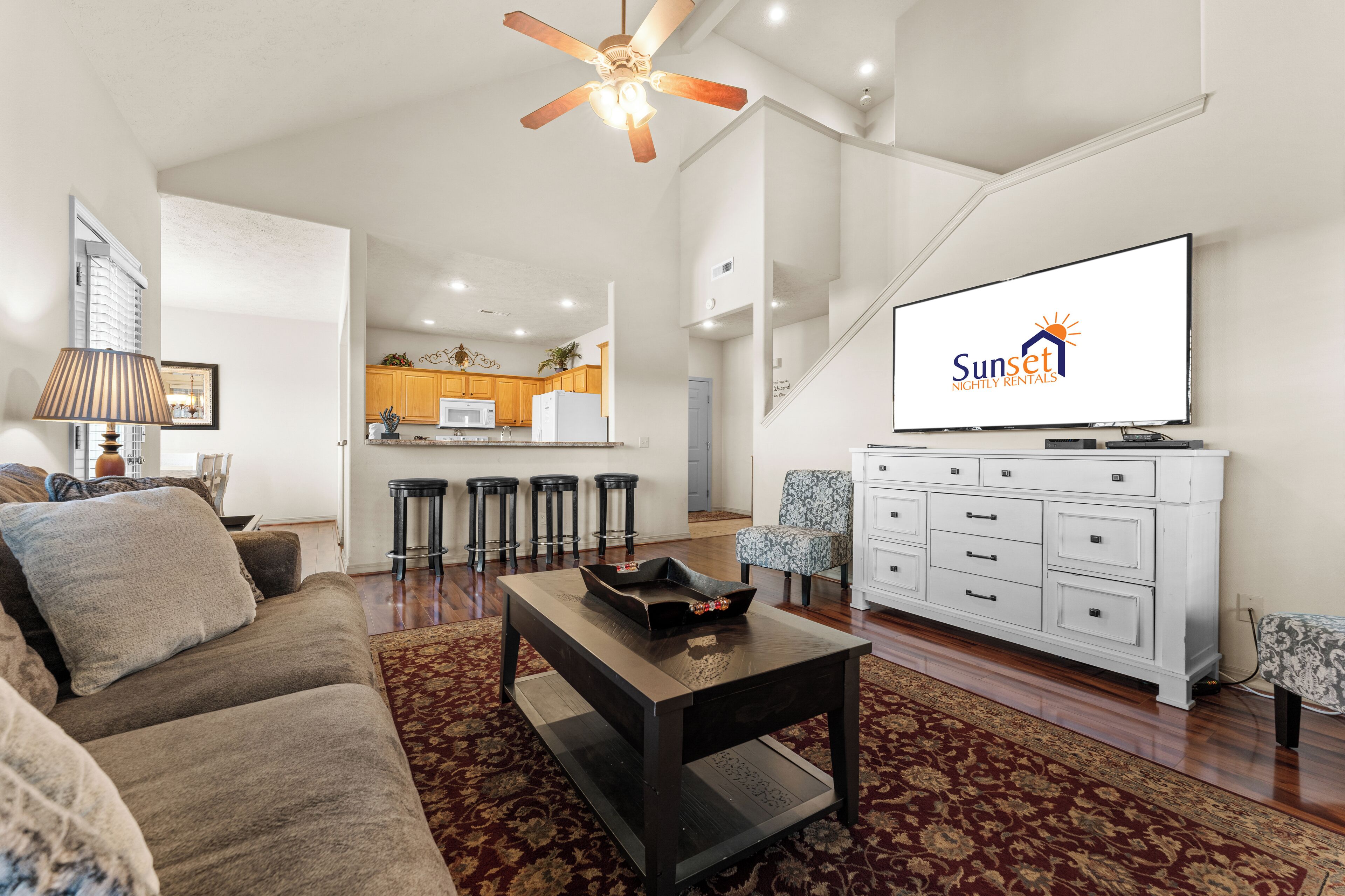 A modern living room with a sofa, coffee table, and TV. There is a kitchen in the background with wooden cabinets and a bar stool area.