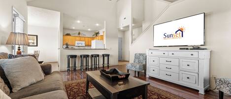 A modern living room with a sofa, coffee table, and TV. There is a kitchen in the background with wooden cabinets and a bar stool area.