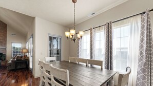 Un espace repas avec une table en bois et des chaises blanches, un lustre et de grandes fenêtres ornées de rideaux à motifs.