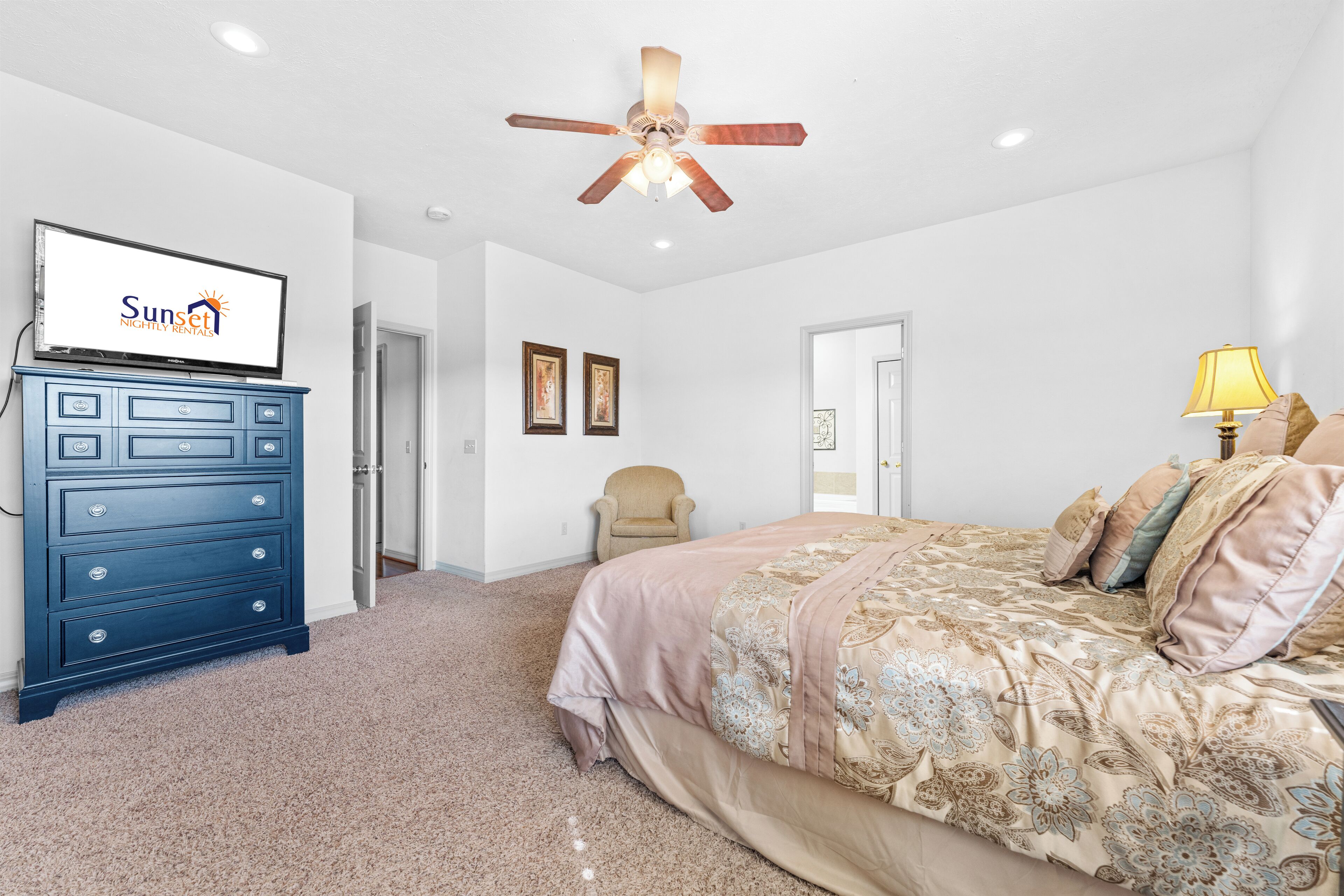 Un dormitorio con cama, ventilador de techo, cómoda, silla y televisor que muestra un logo.