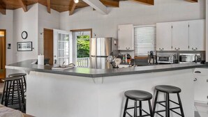 Une cuisine moderne dotée d’un îlot central, d’appareils électroménagers en acier inoxydable et de meubles blancs. L’îlot est agrémenté de deux tabourets de bar, et une grande fenêtre, munie de stores, apporte de la lumière naturelle.
