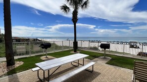 Un patio con una mesa de picnic blanca, dos parrillas y vista a la playa.