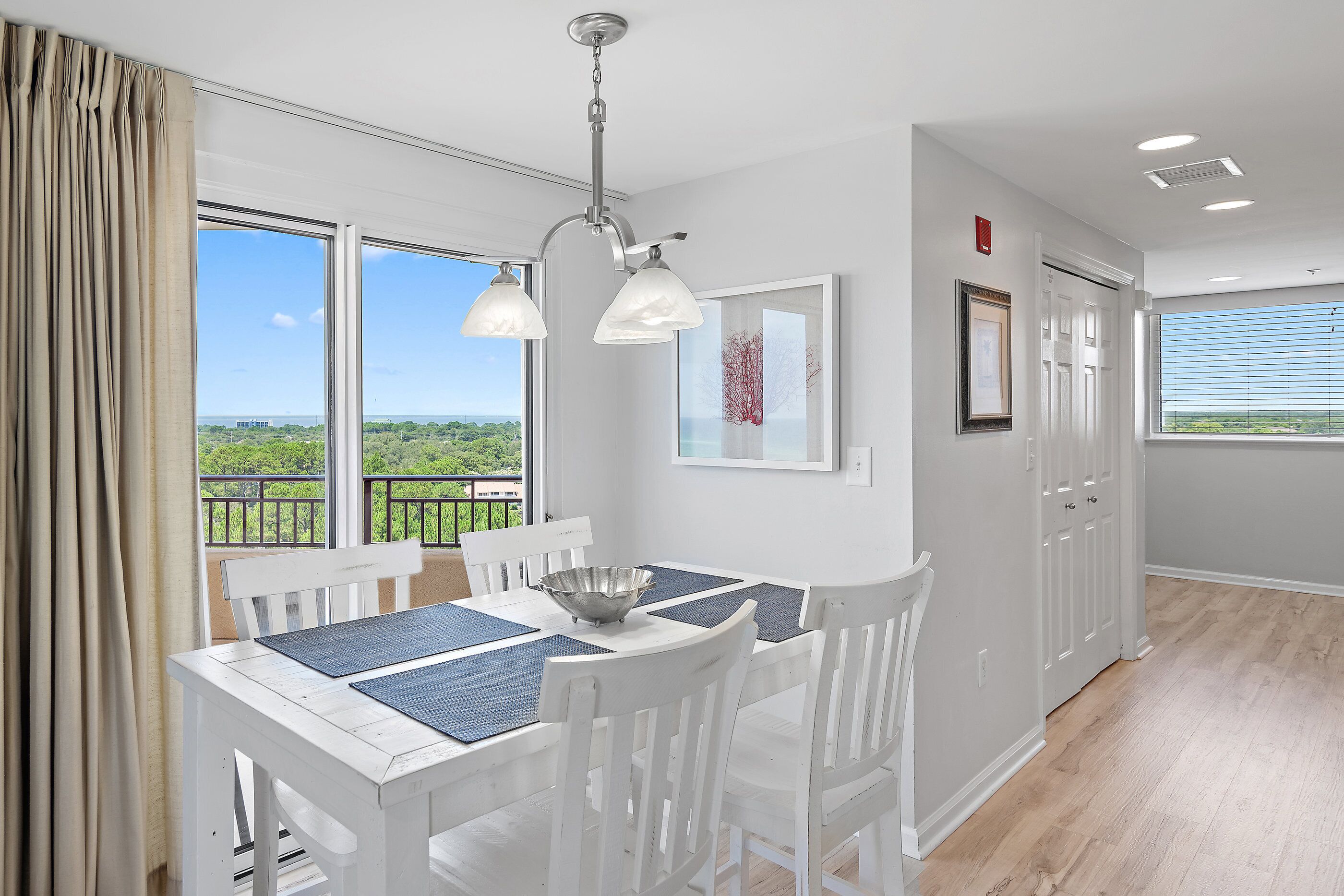 A dining area with a white table and chairs, a hanging light fixture, and large windows offering a view of greenery.
