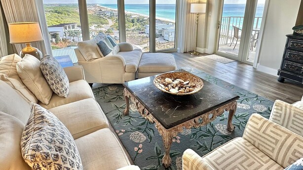 Een ruime woonkamer met een groot raam met uitzicht op het strand en de oceaan. Er is een bank, een salontafel en een lamp.