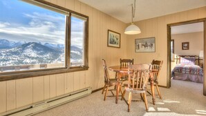 Un coin repas avec une table et des chaises, une grande fenêtre donnant sur les montagnes, et une chambre à coucher avec un lit et un tableau.
