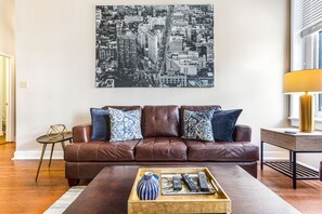 Ein Wohnzimmer mit braunem Ledersofa, gemusterten Kissen, einem Couchtisch mit Fernbedienungen und einem Stadtbild an der Wand.