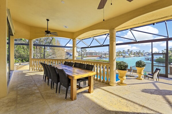 A covered outdoor dining area with a large table and chairs, overlooking a pool and a scenic view.