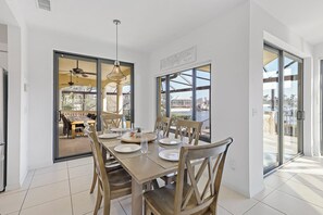 A dining area with a large table set for six, a ceiling fan, and large windows offering a view of the outdoors. - Serene Waterfront Home | Pool, Spa, & Water View (Marco Island)