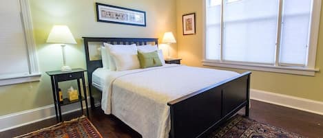 A bedroom with a bed, two bedside tables with lamps, a window with blinds, and a patterned rug.