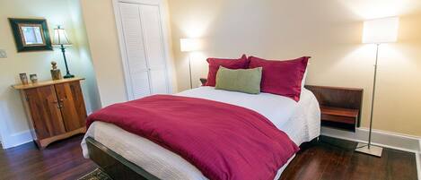 A bedroom with a large bed, a wooden nightstand, a bedside table, a floor lamp, and a door.