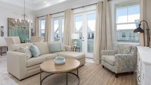 A spacious living room with a beige sofa, patterned armchair, and a round coffee table. There is a large window with curtains, a chandelier, and a lamp on a side table. - Luxurious Townhouse on 30a! 6-Seat LSV, Beach, Pool, Walk to Shopping+Dining. (Panama City)