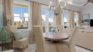 A spacious living room with a round table, beige upholstered chairs, a chandelier, and a large window with curtains. - Luxurious Townhouse on 30a! 6-Seat LSV, Beach, Pool, Walk to Shopping+Dining. (Panama City)