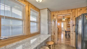 Eine Küche mit Holzausstattung, einer Arbeitsplatte aus Granit und einem Kühlschrank. Zwei Holzbarhocker und ein Fenster mit Jalousien sind vorhanden.