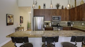 A modern kitchen with granite countertops, stainless steel appliances, and wooden cabinets. There are three pendant lights, a refrigerator, a microwave, and a stove.