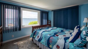 Une chambre avec une grande fenêtre, un lit recouvert d’une couette bleue et blanche, et une vue sur l’eau.