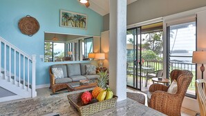 A spacious living area with a ceiling fan, a large window offering a view of the outdoors, a dining area with wicker furniture, and a fruit bowl on the table.