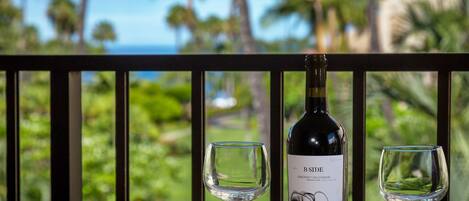 Una botella de vino y dos copas sobre una mesa, con vista a palmeras y un cuerpo de agua.