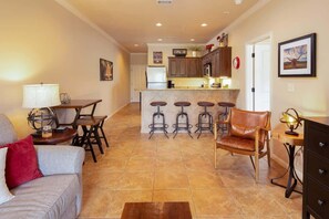 A living room with a sofa, a coffee table, a dining table with chairs, a kitchen area with a refrigerator, and a bar with stools.