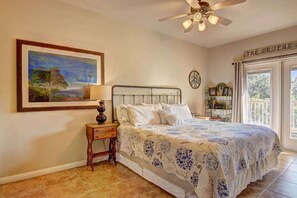 A bedroom with a bed, a nightstand, a painting, a ceiling fan, and a window with a view.
