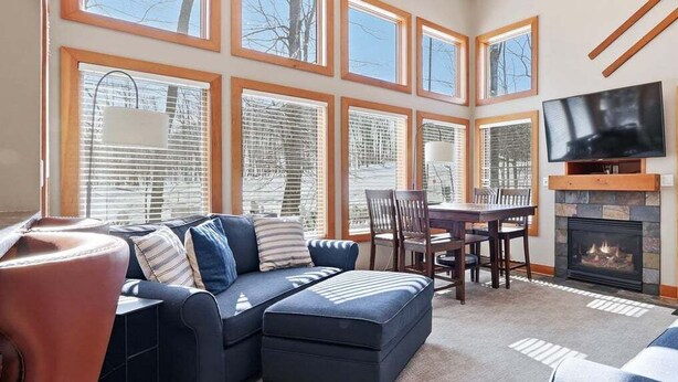 A spacious living room with a fireplace, a television mounted above it, and a dining area with a table and chairs. - Newly Remodeled 780 Disciples Condo | Slopeside, short walk to Village Lane (Boyne Falls)