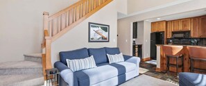 A living room with a blue sofa, a wooden staircase, and a kitchen area with wooden cabinets and appliances. - Newly Remodeled 780 Disciples Condo | Slopeside, short walk to Village Lane (Boyne Falls)