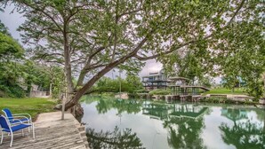 A wooden deck with blue chairs, a pond, and a building with a bridge. - LD 1683 Texas River Haus (New Braunfels)
