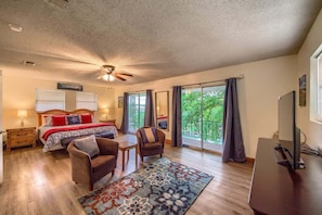 A bedroom with a bed, two chairs, a desk, a ceiling fan, and large windows with curtains.