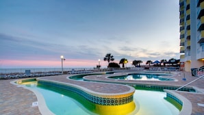 Un área de piscina con múltiples piscinas y sillones de descanso, rodeada por una cerca y palmeras.
