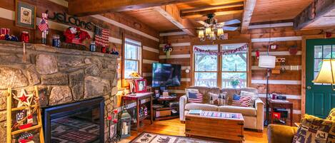 A cabin interior with a stone fireplace, a television, a couch, and a sign reading "Cabin Sweet Cabin".