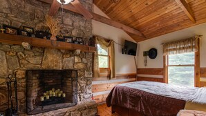 Une pièce rustique avec une cheminée en pierre, des poutres apparentes, un lit recouvert d’une couverture marron et une fenêtre agrémentée de rideaux.