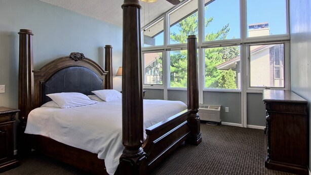A four-poster bed with a wooden headboard, a ceiling fan, and large windows offering a view of the outdoors. - Newly Renovated 4BR / 4BA Pet-Friendly, Best Walking Location At Boyne Mountain (Boyne Falls)