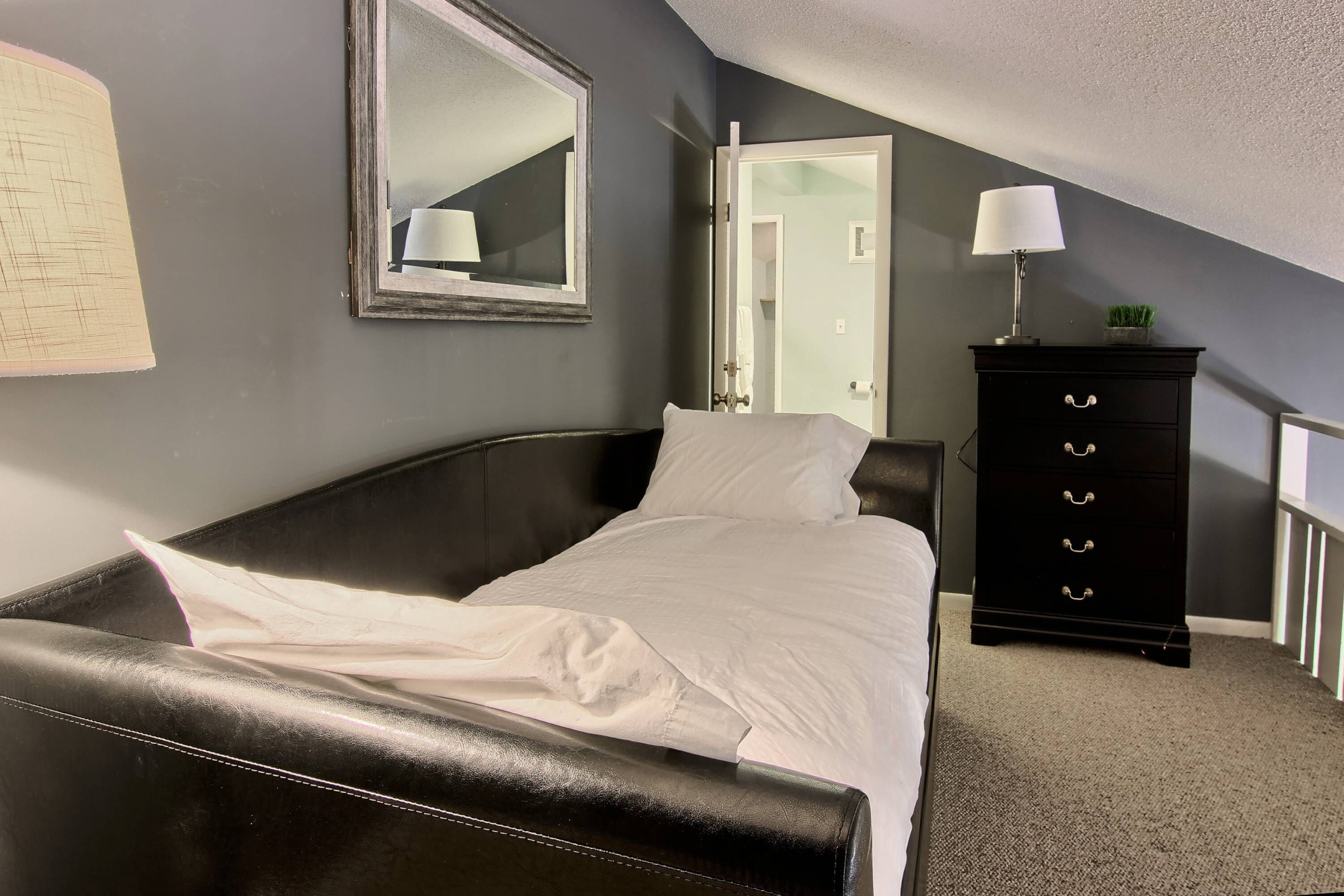 Un dormitorio con una cama de cuero negro, una almohada blanca, un aparador negro con cajones y un espejo en la pared.