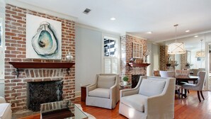 A living room with a brick fireplace, two armchairs, a glass coffee table, and a dining area with a chandelier. - Historic 2 Story Home w/ Balcony, Parking on Calhoun Square by Lucky Savannah (Savannah)