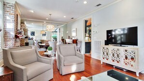 A modern living room with two armchairs, a TV on a wooden cabinet, and a dining area in the background. - Historic 2 Story Home w/ Balcony, Parking on Calhoun Square by Lucky Savannah (Savannah)