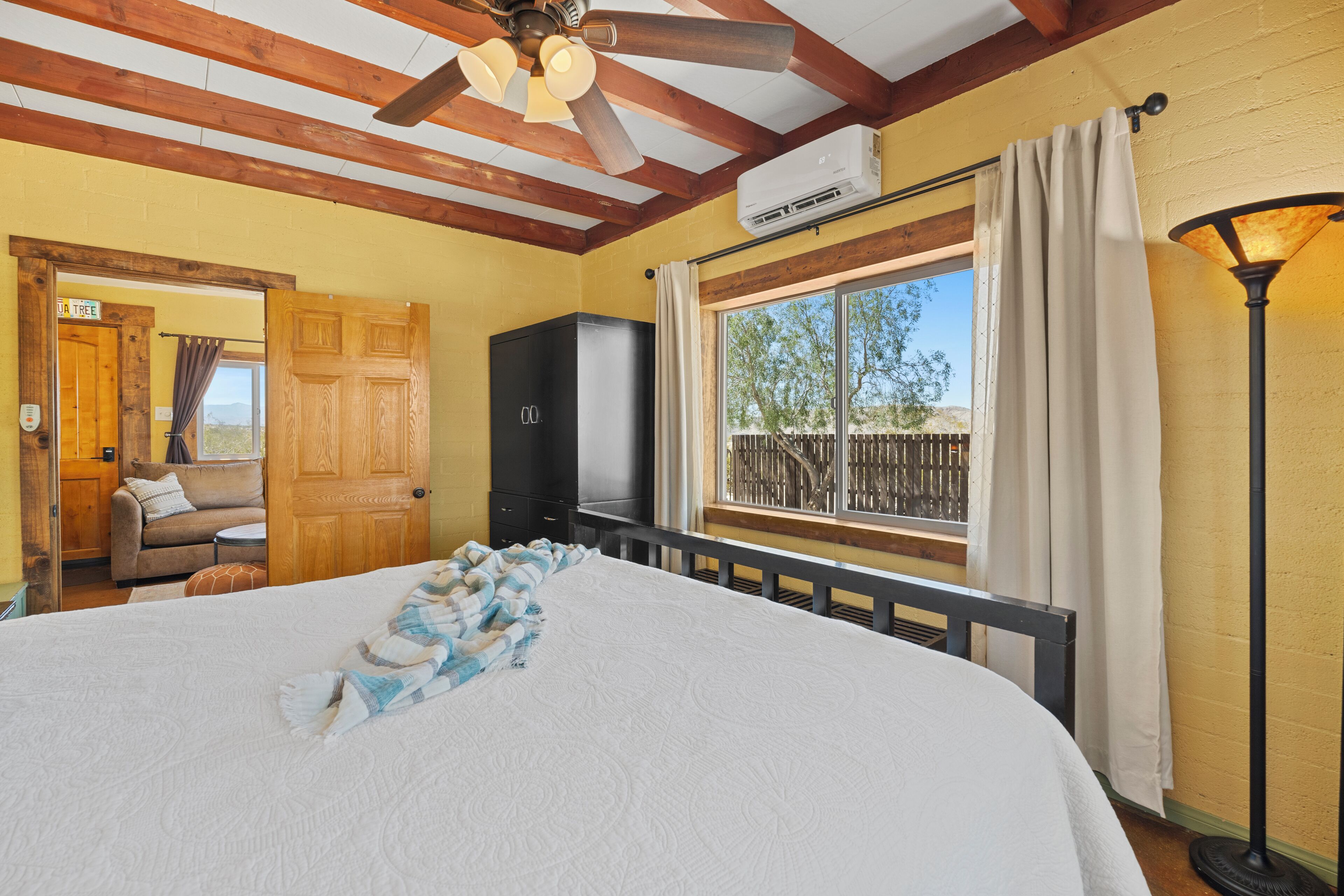 Ein Schlafzimmer mit einem Bett, einem Deckenventilator, einer Klimaanlage, einem Fenster mit Vorhängen und Blick ins Freie.