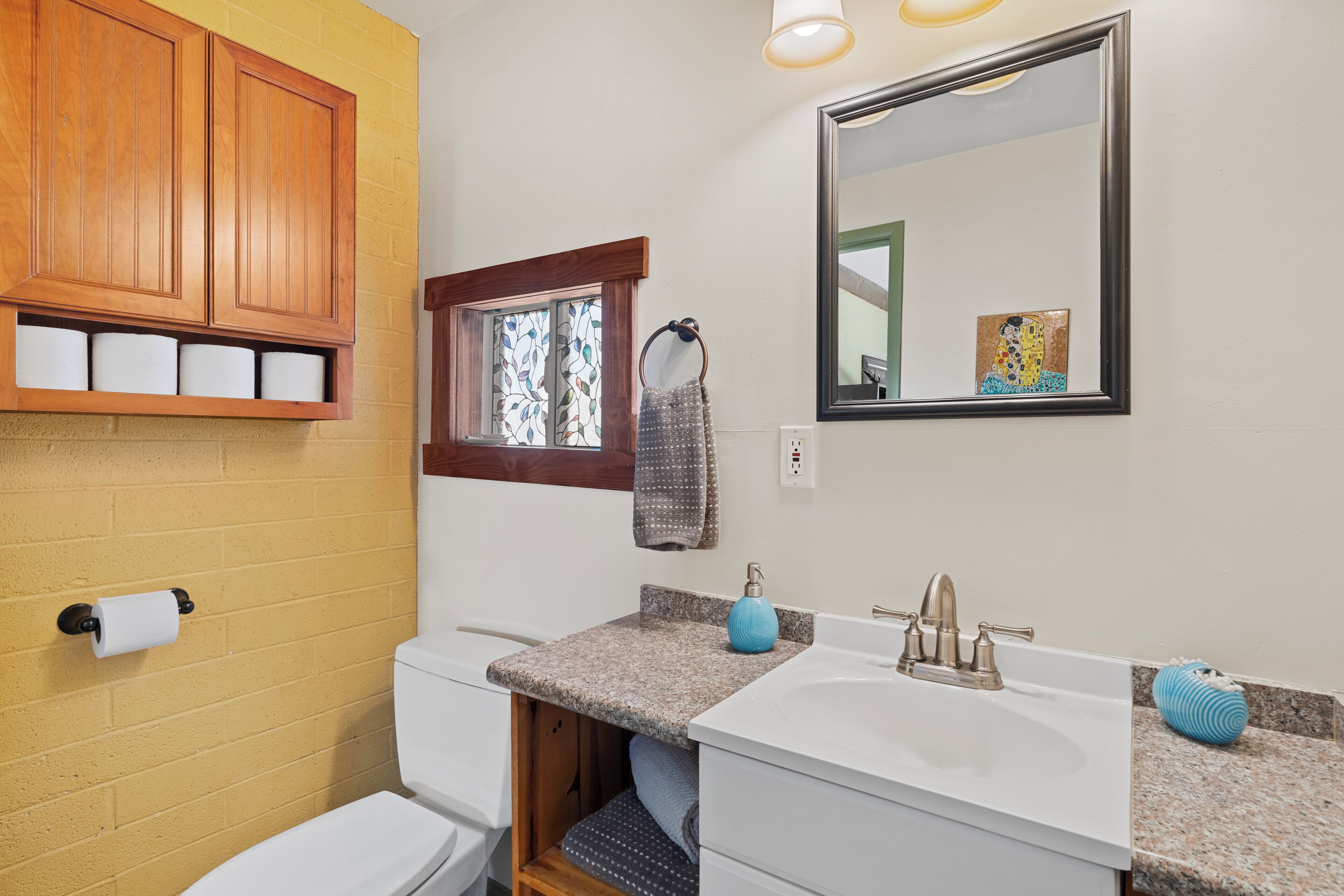 Une salle de bain avec des meubles en bois, des toilettes, un lavabo avec un miroir et une fenêtre où est accrochée une serviette.