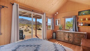 A bedroom with a large bed, wooden walls, a window with a view of the desert, and a wooden dresser with drawers.