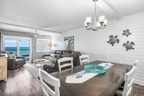 Amplia sala de estar con una mesa de comedor de madera oscura, sillas blancas y vista al océano a través de grandes puertas correderas de vidrio.