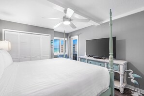 Un dormitorio con una cama grande, un ventilador de techo, una televisión y vista al océano a través de una ventana.