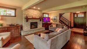 A spacious living room with a stone fireplace, a television, a staircase, and a large window. - Walk to the Winery! On the Golf Course in Suncadia! Hot Tub | Up to 33% Off!! (Cle Elum)