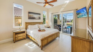 A bedroom with a large bed, a ceiling fan, a nightstand, a lamp, a painting, a TV, and a view of an outdoor pool area. - Waterfront Home | Pool, Spa & Walk to Beach (Marco Island)