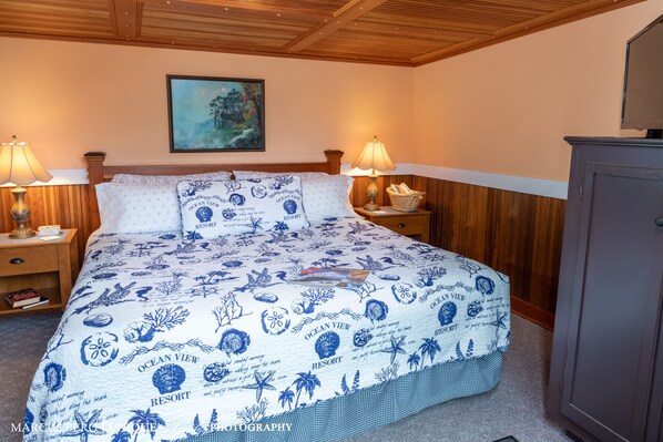 A bedroom with a bed, two bedside tables, lamps, a framed painting, and a wooden cabinet.