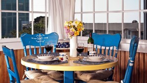 A sunlit room with a round table set for two, featuring blue chairs with white text, a vase of flowers, and a bottle of wine.