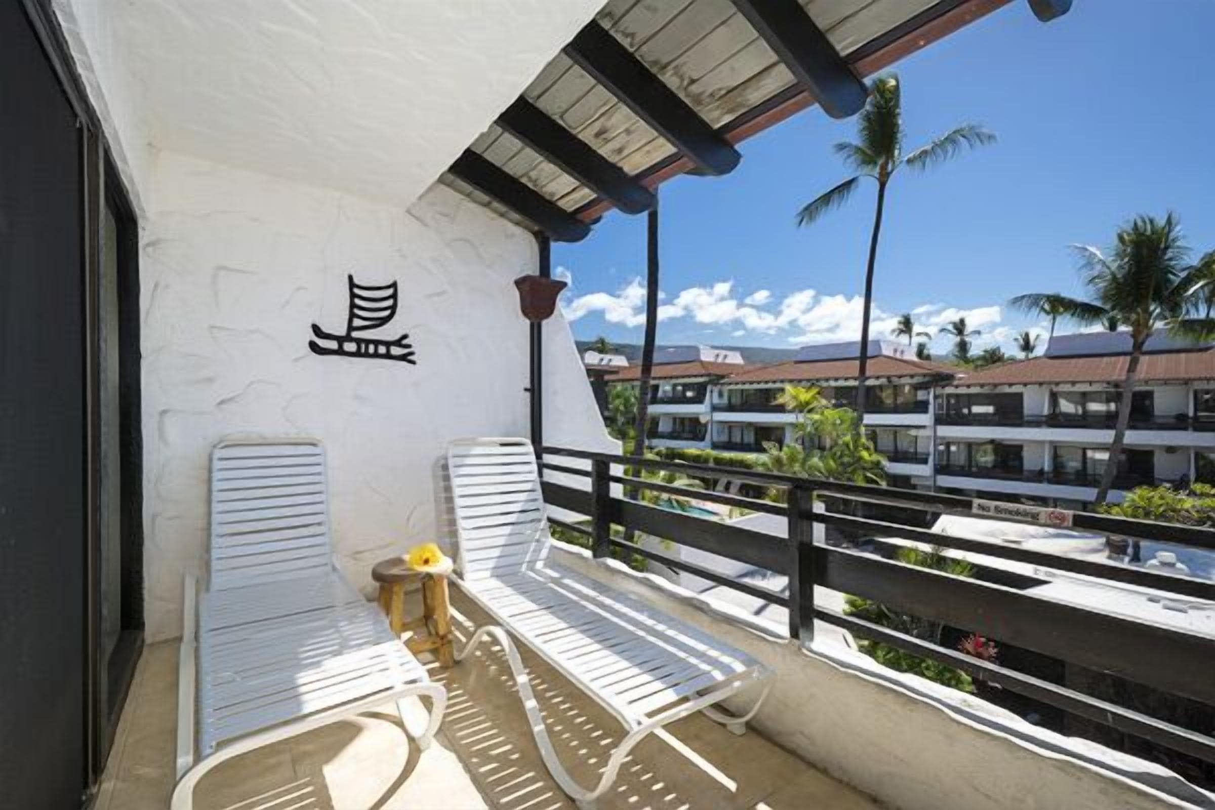 Un balcón con sillones blancos, una mesita con un florero y vistas a una zona residencial con palmeras y casas.
