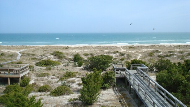 Ein Strand mit einem hölzernen Promenadenweg, einem sandigen Pfad und spärlicher Vegetation.